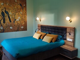 A bedroom with a large bed, two bedside tables, a wall-mounted light fixture, and a painting of birds on a tree.