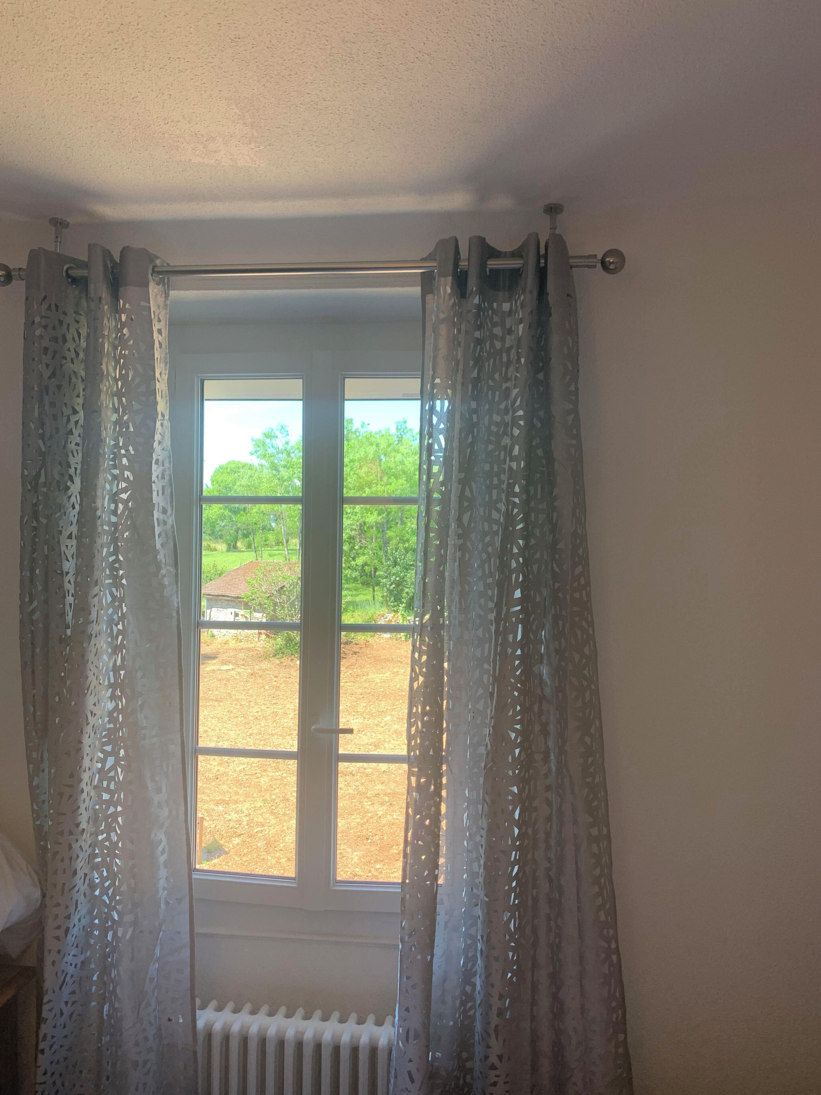A room with a window featuring patterned curtains, a radiator below, and a view of a green landscape outside.