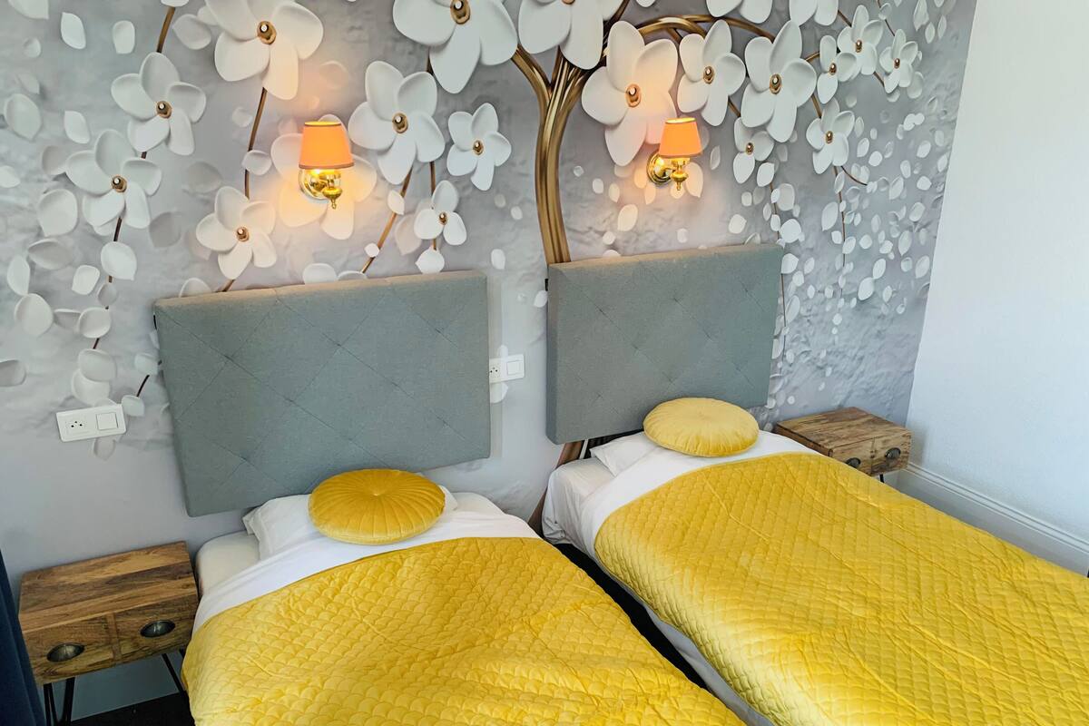Een kamer met twee bedden, elk met een hoofdeinde en een gequilt, geel sprei. Er staan wandlampen met oranje kapjes en de muur is versierd met witte bloemenpatronen.