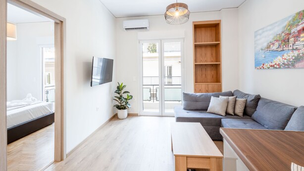 A modern living room with a grey sofa, a wooden coffee table, a potted plant, and a TV mounted on the wall. - Modern 2-bedroom Gem Close to Metro (Athens)