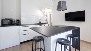 Ett modernt kök med vita skåp, en svart bänkskiva och en stor köksö med barstolar. En platt-TV är monterad på väggen ovanför köksön.