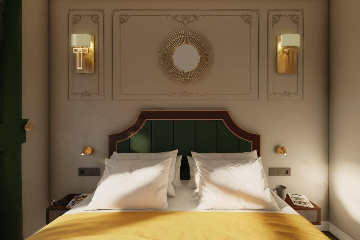 A neatly made bed with white linens and a green headboard, two bedside lamps, a wall-mounted mirror, and a decorative wall piece.