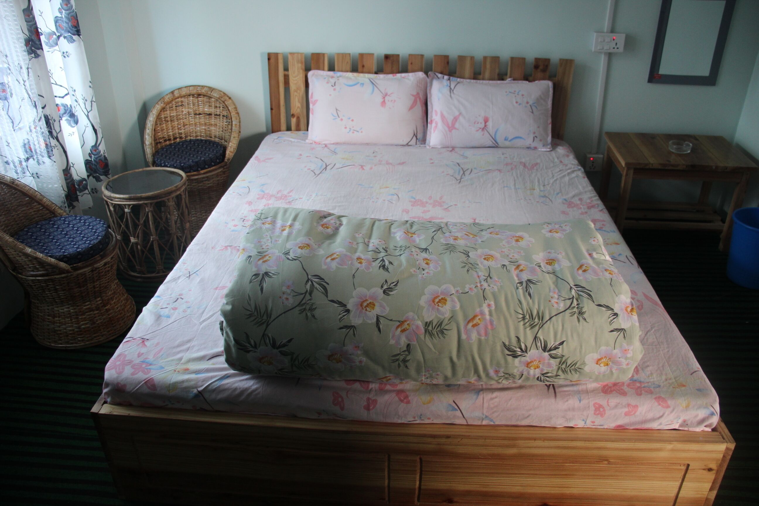 A bed with floral bedding, two wicker chairs, a small round table, and a nightstand with a lamp.