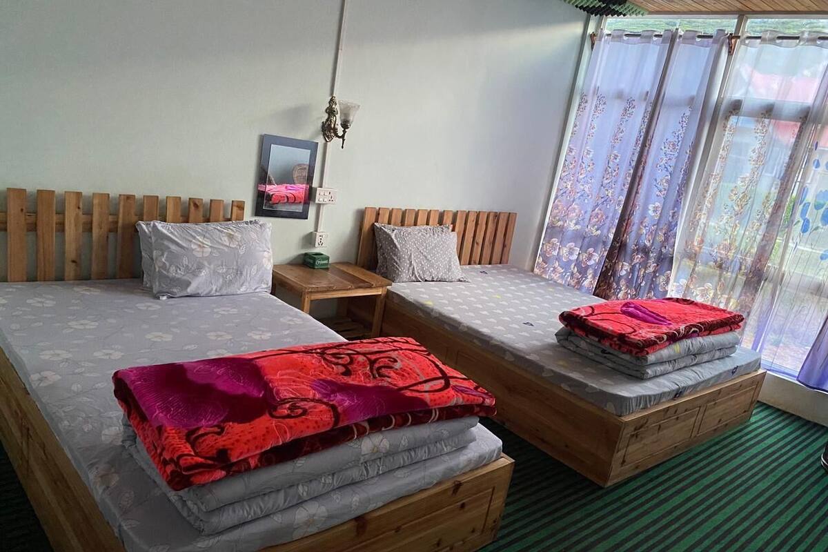 Une chambre avec deux lits en bois, chacun étant équipé d’un matelas et d’une couverture rouge. On y trouve une table de chevet avec une lampe, une tête de lit en bois et une fenêtre avec des rideaux.