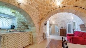 Habitación con paredes de piedra, una zona de cocina, un sofá rojo y piso de baldosas.