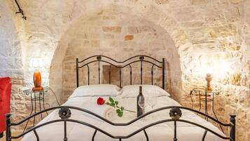 Un lit avec un cadre en métal, une literie blanche, une rose rouge, une bouteille et des verres, le tout placé devant un mur en pierre.