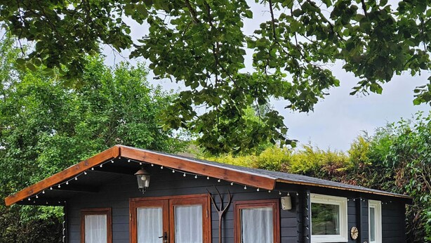 A small, dark-colored cabin with a sloped roof, two wooden doors, and a small porch area with a lantern and a potted plant. - The AleXan balneotherapy chalet (Quinssaines)