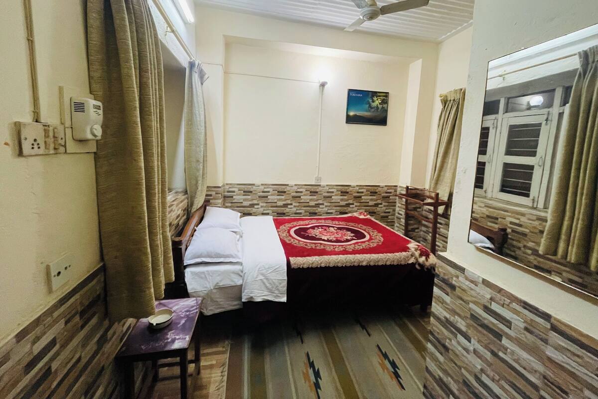 A small, well-lit room with a bed, a ceiling fan, a window with curtains, a wall-mounted air conditioner, and a small table with a bowl.