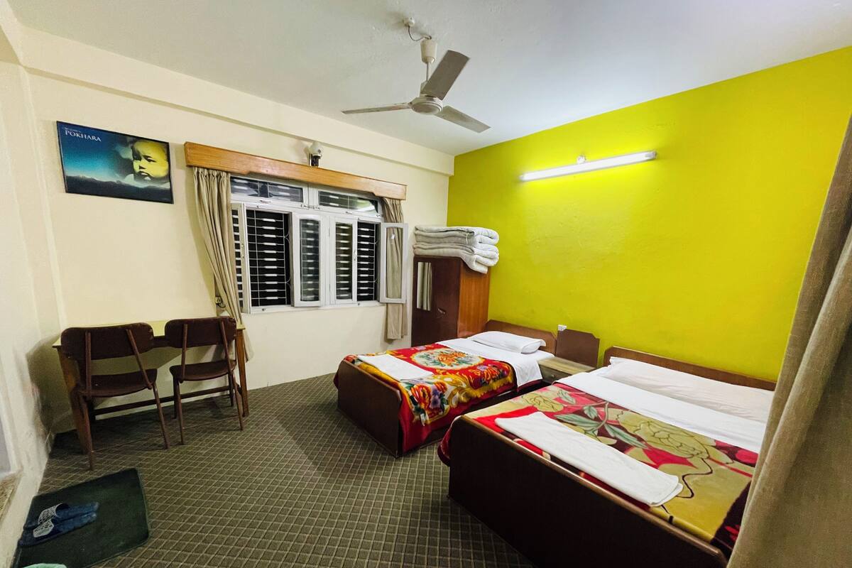 A hotel room with two beds, a desk, a chair, a ceiling fan, and a window with shutters.