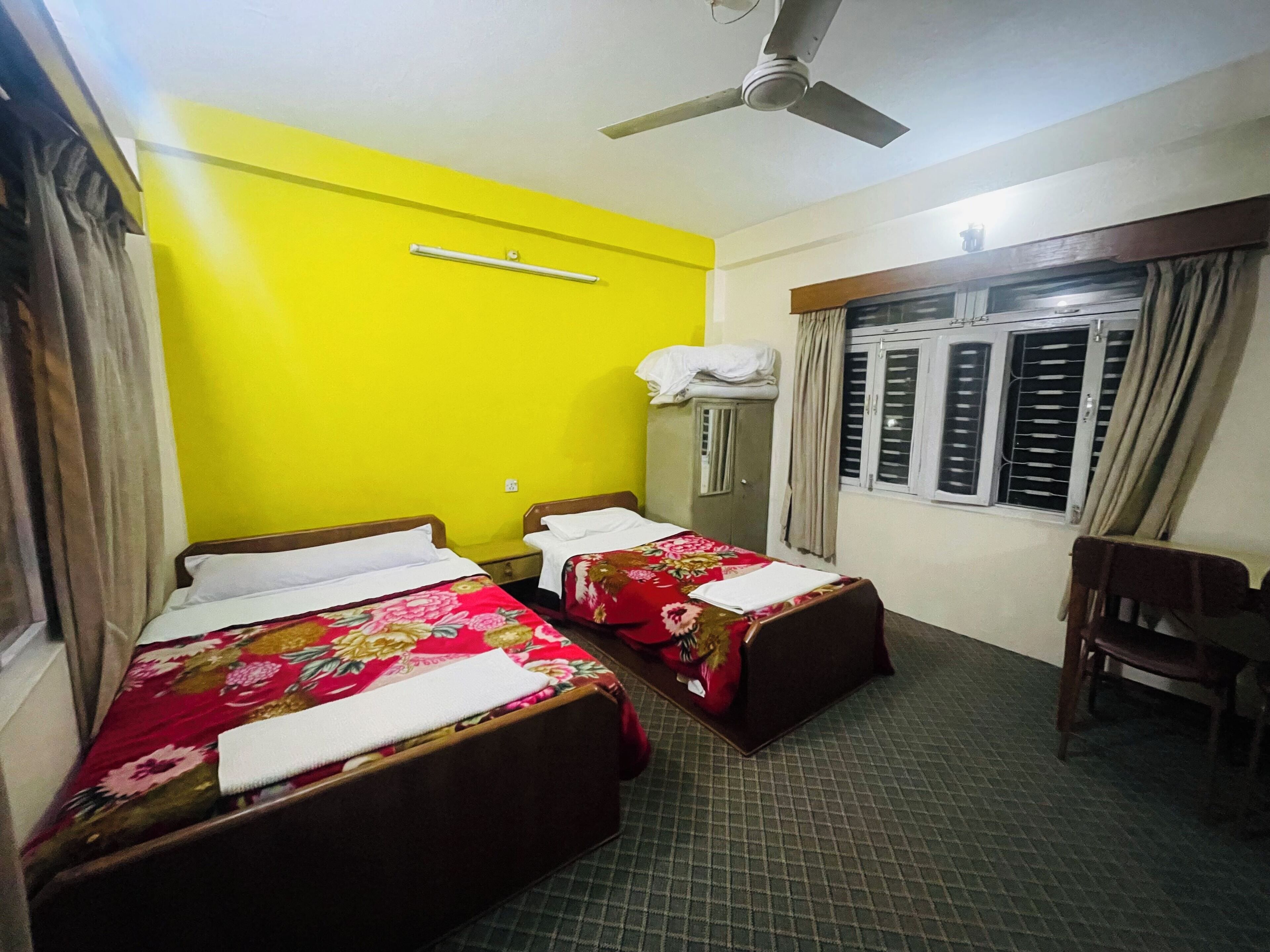 Une chambre d’hôtel avec deux lits, un ventilateur de plafond, une fenêtre avec des rideaux et un petit bureau avec une chaise.