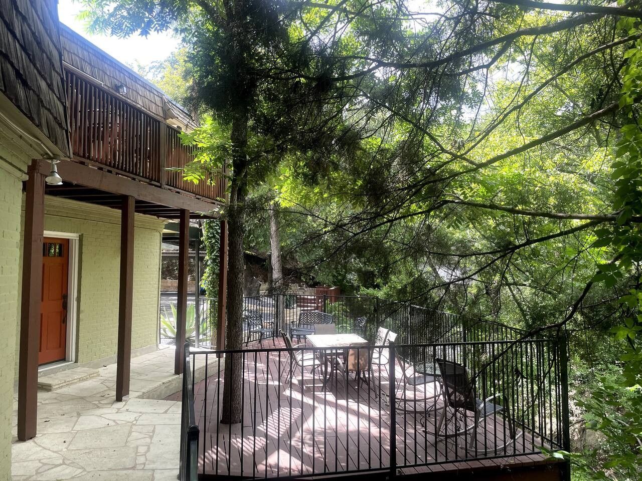 A covered outdoor patio with a table and chairs, surrounded by a metal fence and trees.