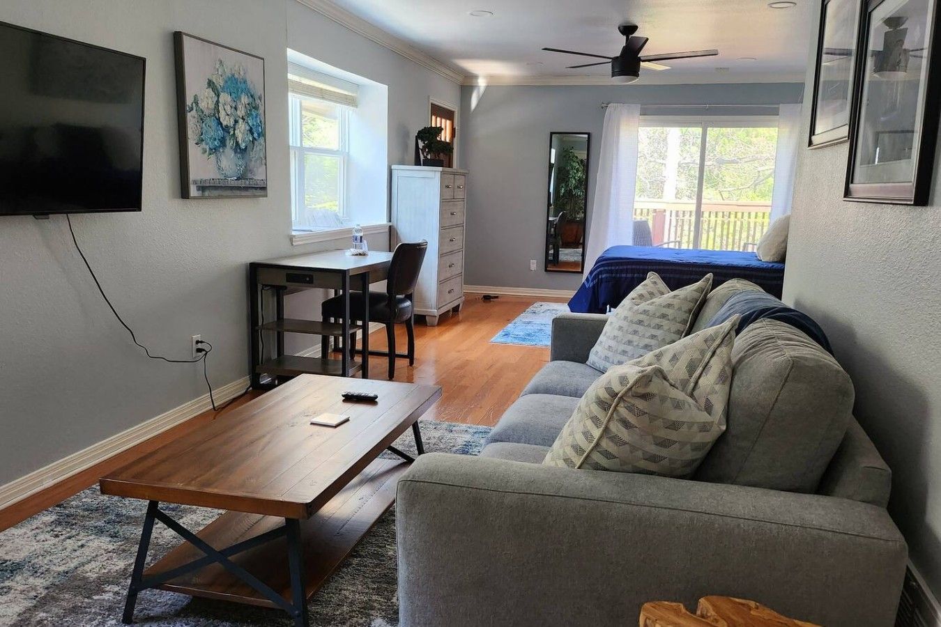 A living room with a sofa, a coffee table, a TV, a desk, and a chair. There is a window with curtains and a ceiling fan.