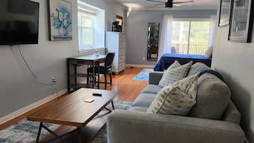 A living room with a sofa, a coffee table, a TV, a desk, and a chair. There is a window with curtains and a ceiling fan.