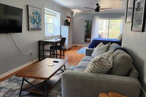 A living room with a sofa, a coffee table, a TV, a desk, and a chair. There is a window with curtains and a ceiling fan. - Travis Heights Oasis! (Austin)