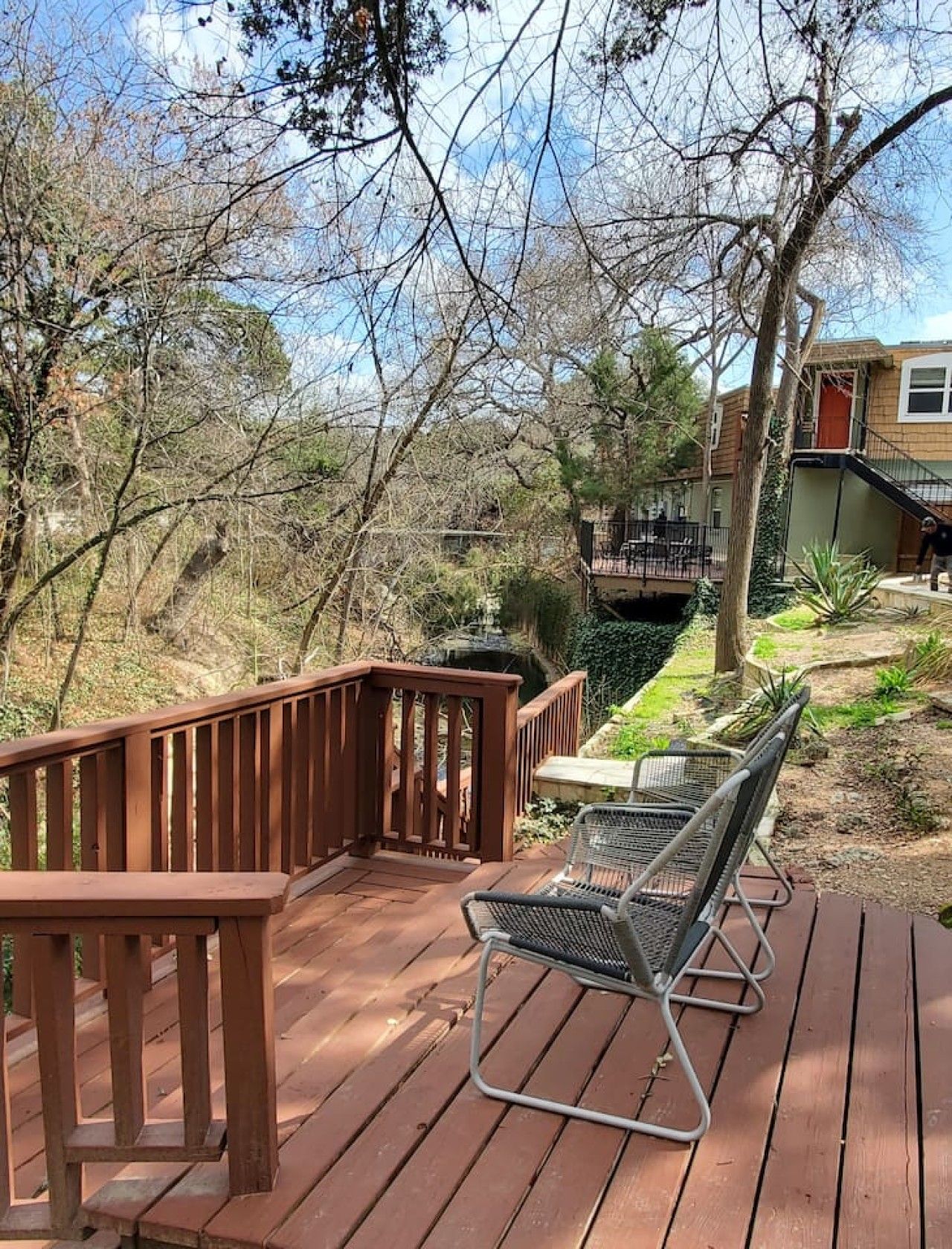 A wooden deck with a metal chair, surrounded by trees and a small bridge.