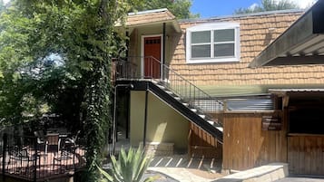 A two-story house with a wooden exterior, a front door, a window, a staircase, and a patio area with chairs.