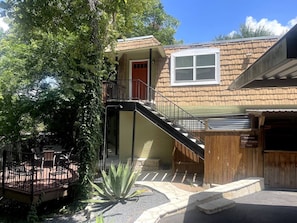 A two-story house with a wooden exterior, a front door, a window, a staircase, and a patio area with chairs.