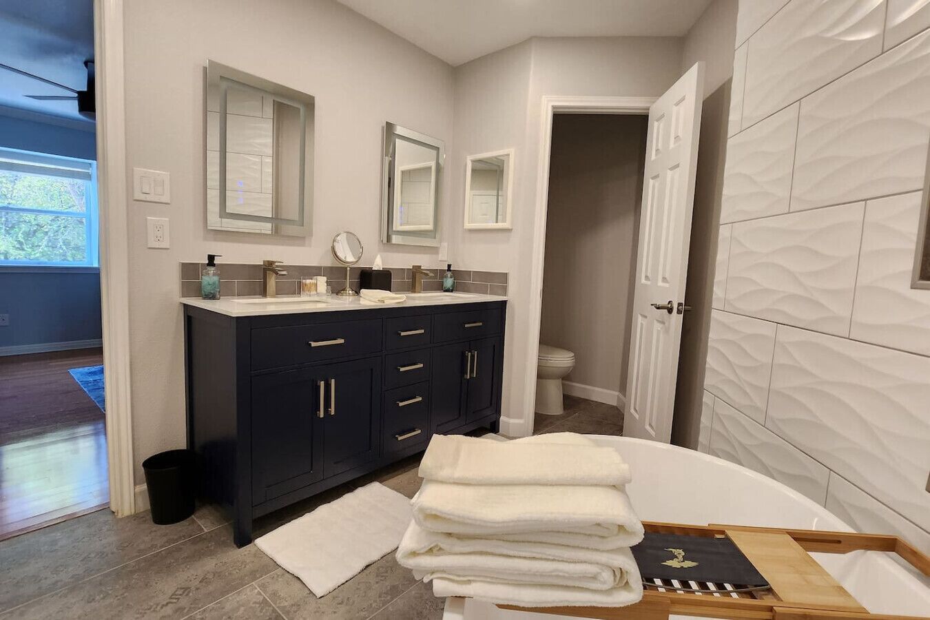 A modern bathroom with dark cabinets, a white sink, and a large mirror. There is a stack of towels on a tray and a wooden bench.
