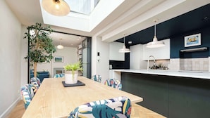 Uma área de refeições moderna com uma mesa de madeira comprida, cadeiras coloridas, um vaso de planta e iluminação pendente.