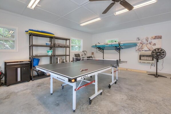 A ping pong table in a room with a ceiling fan, shelves, and a surfboard.