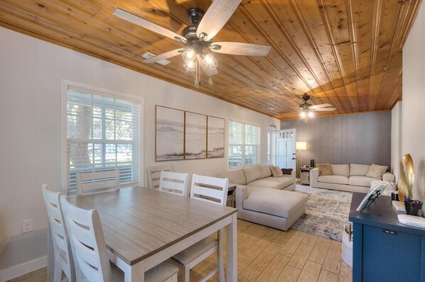 A spacious living area with a wooden ceiling, a dining table with chairs, a sofa, and a small side table with a lamp.