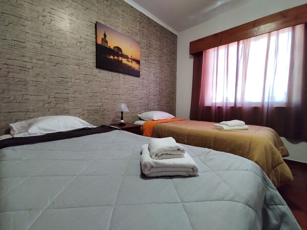 A hotel room with two beds, a brick wall, a painting, a window with curtains, and a nightstand with a lamp.