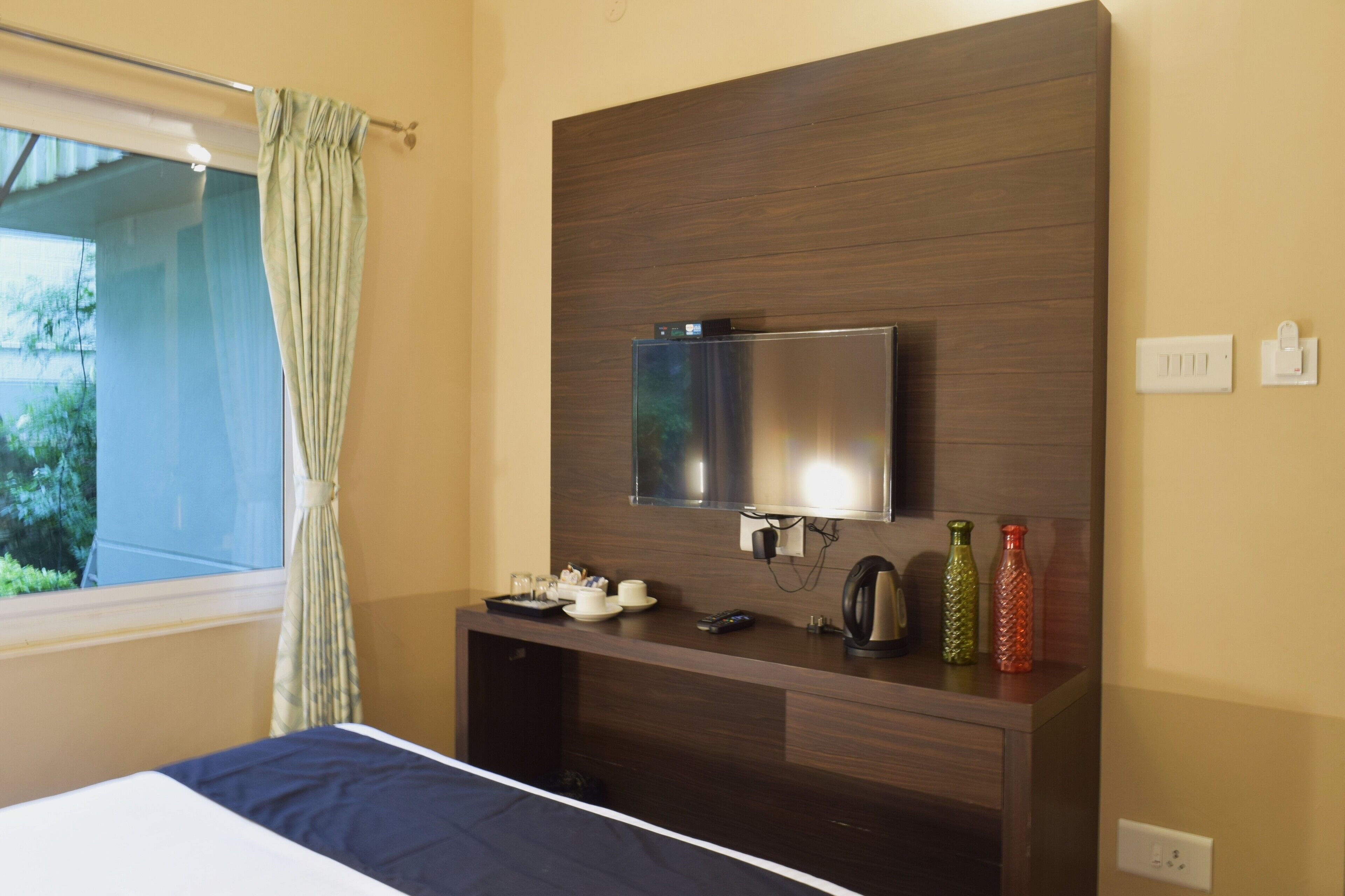 A hotel room with a bed, a TV mounted on a wooden panel, a desk with a kettle, and a window with curtains.