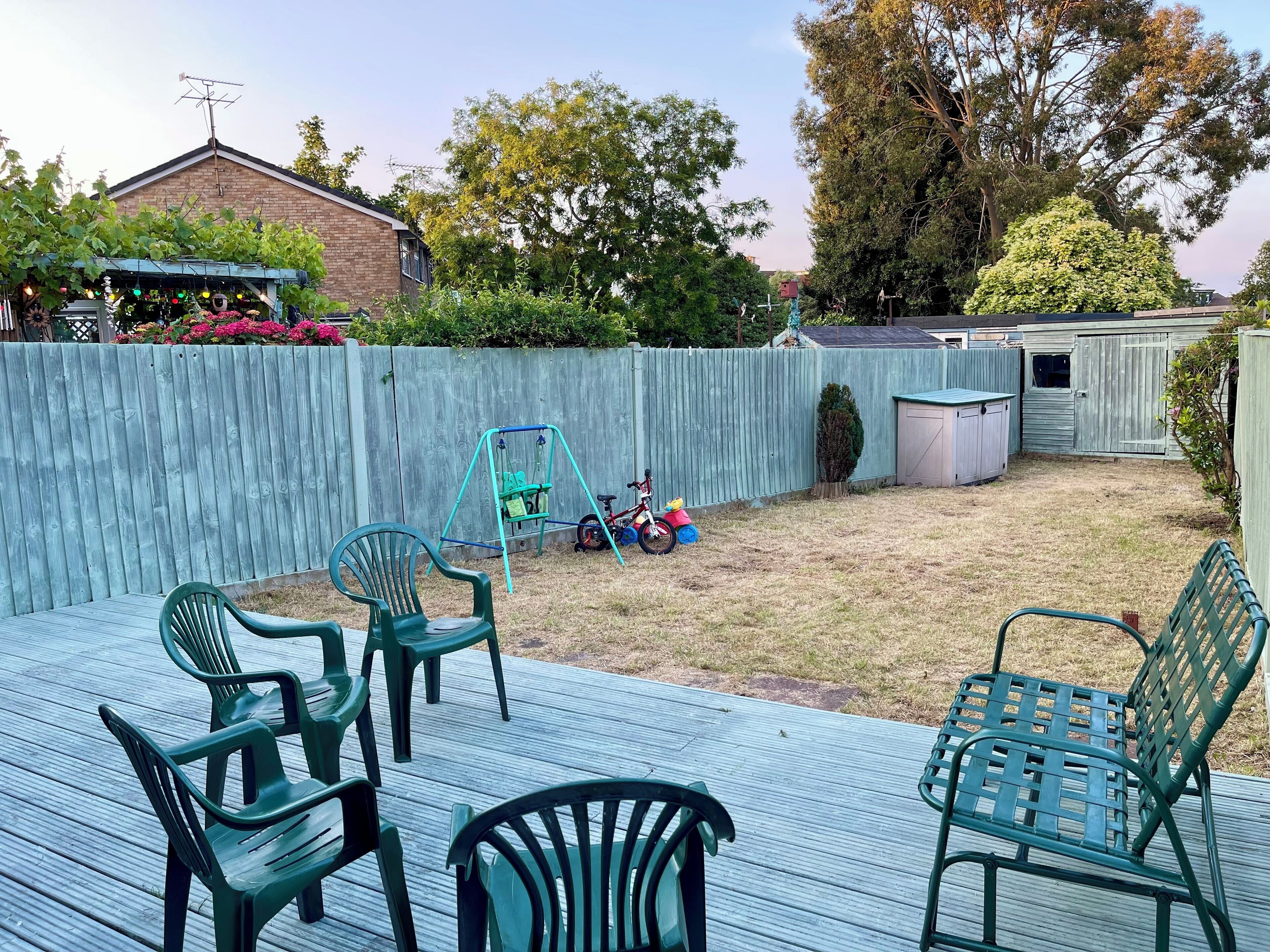 Een achtertuin met een houten terras, groene plastic stoelen, een schommel en een tuinhuis.