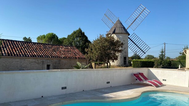 A swimming pool with a stone wall, a windmill, and a building with a tiled roof.
