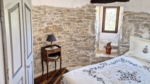 A bedroom with a stone wall, a wooden nightstand with a lamp, a window with a view, and a bed with a floral bedspread.