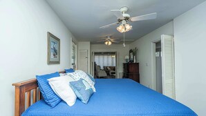 Une chambre avec un grand lit, un ventilateur de plafond et une porte donnant sur une autre pièce.