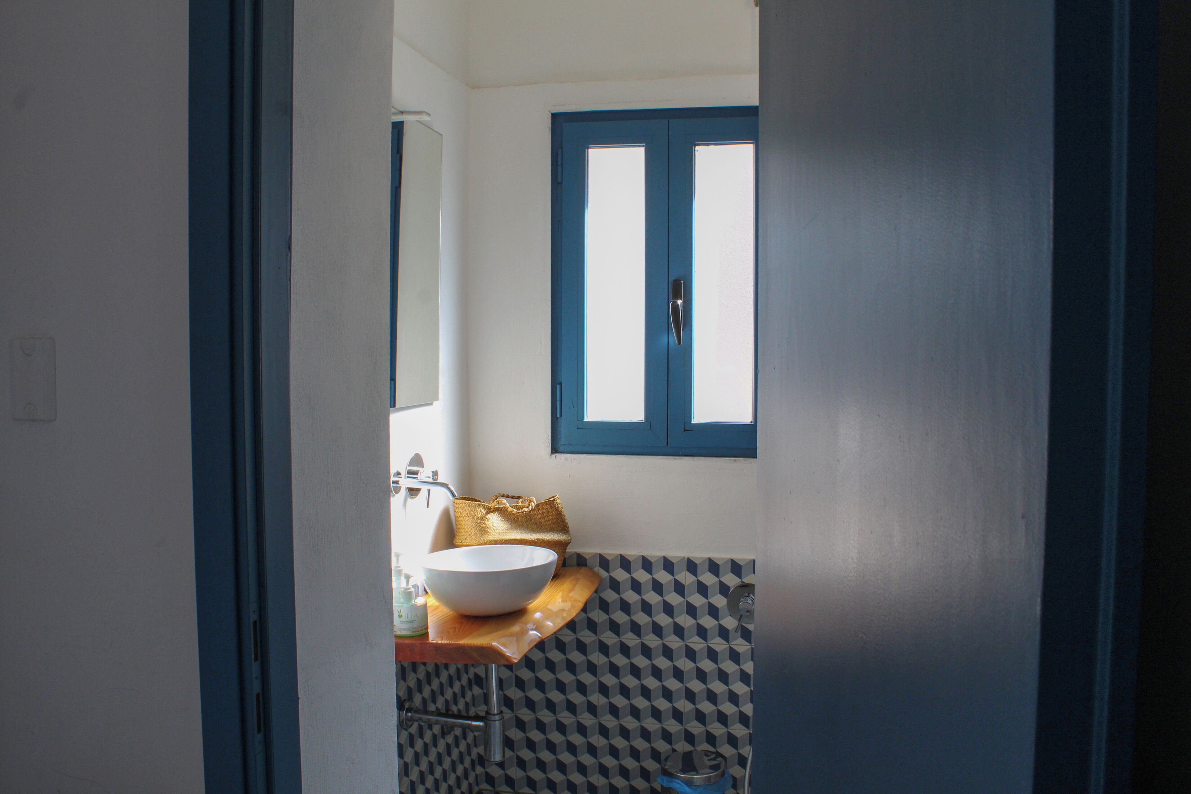 Un baño con un lavamanos blanco, un bolso dorado y una ventana con marco azul.
