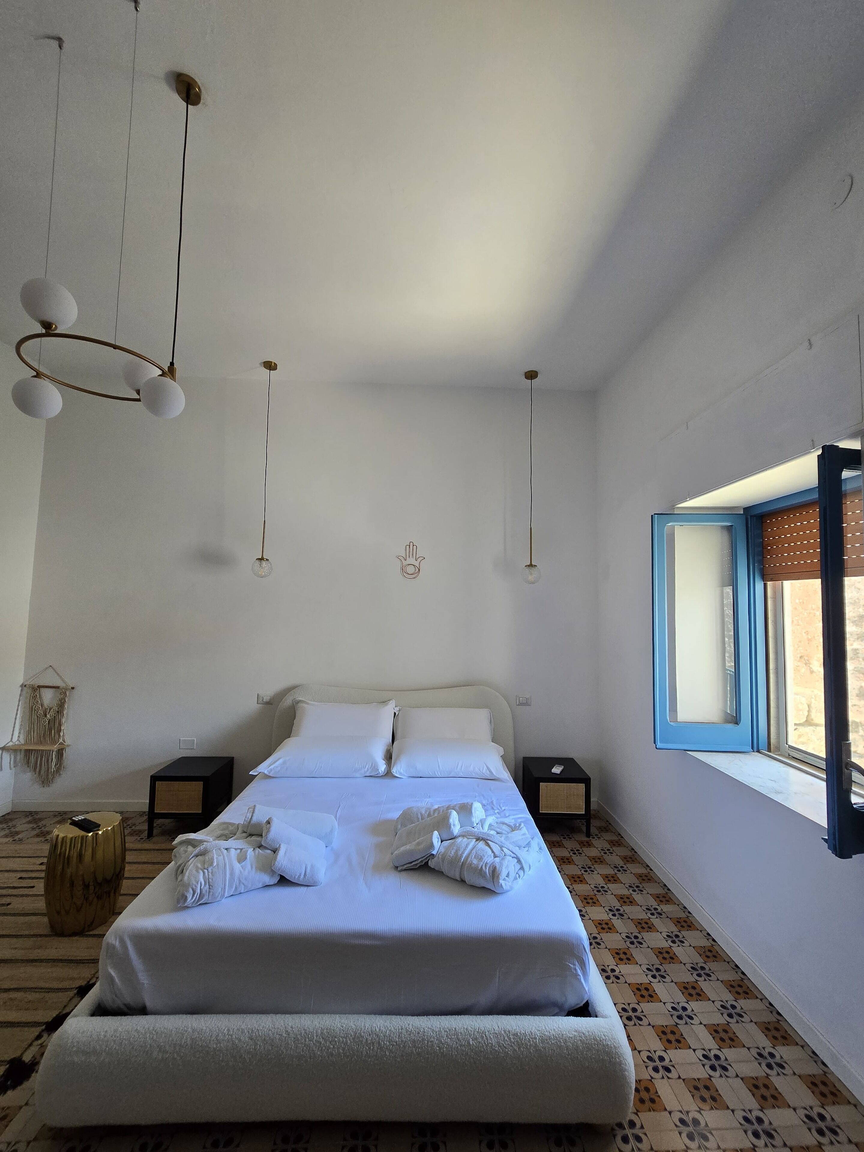 Una cama bien tendida con sábanas blancas, una lámpara colgante y una ventana con contraventanas azules.