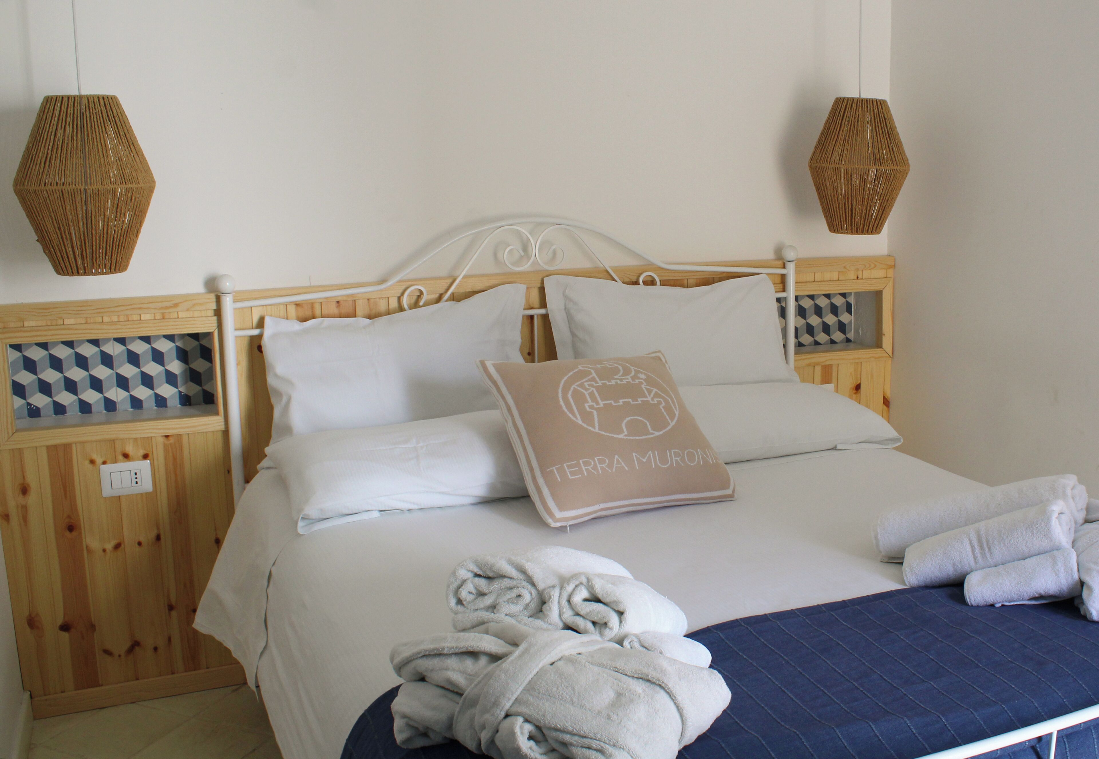 A neatly made bed with white linens, a pillow with 'TERRA MURONI' text, and two woven wall lamps.