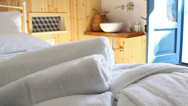 Una stanza con pareti in legno, un letto con lenzuola bianche, un comodino in legno con cuscino a quadri, uno scaffale in legno con un lavabo bianco e articoli da toilette, e una porta che dà sull'esterno.