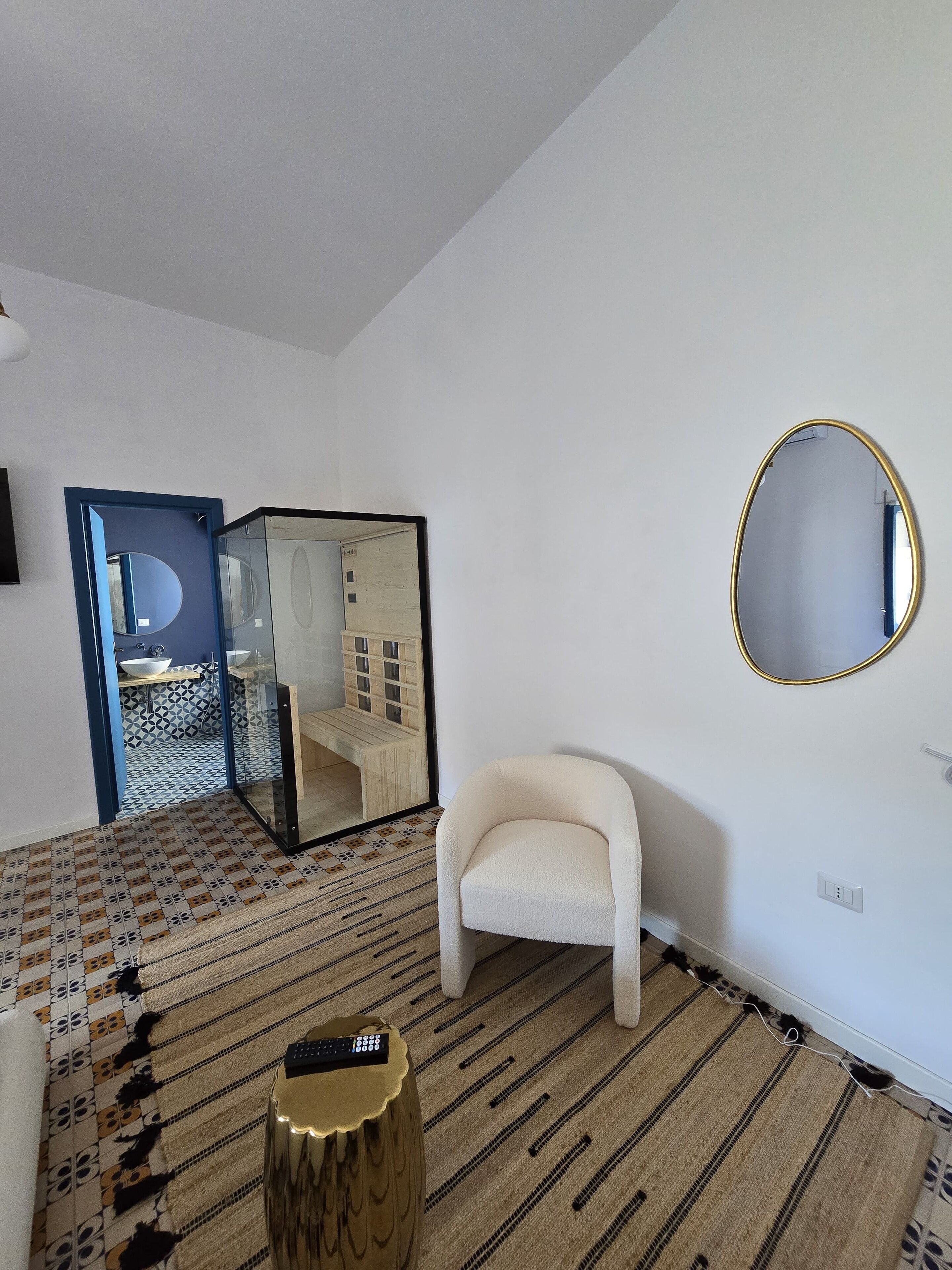 Una habitación con un sillón blanco, un espejo redondo y un taburete de madera con un control remoto.