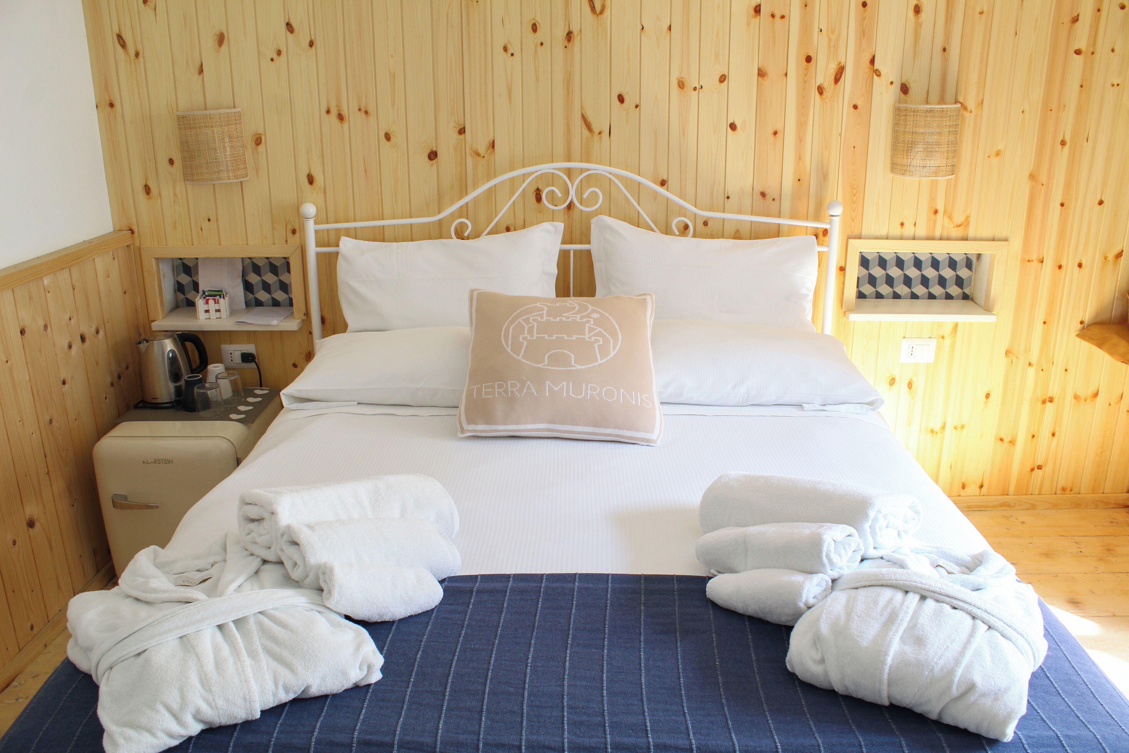 A neatly made bed with white linens, a blue striped bedspread, and a pillow with a logo. There are towels and a small table with a kettle and other items beside the bed.