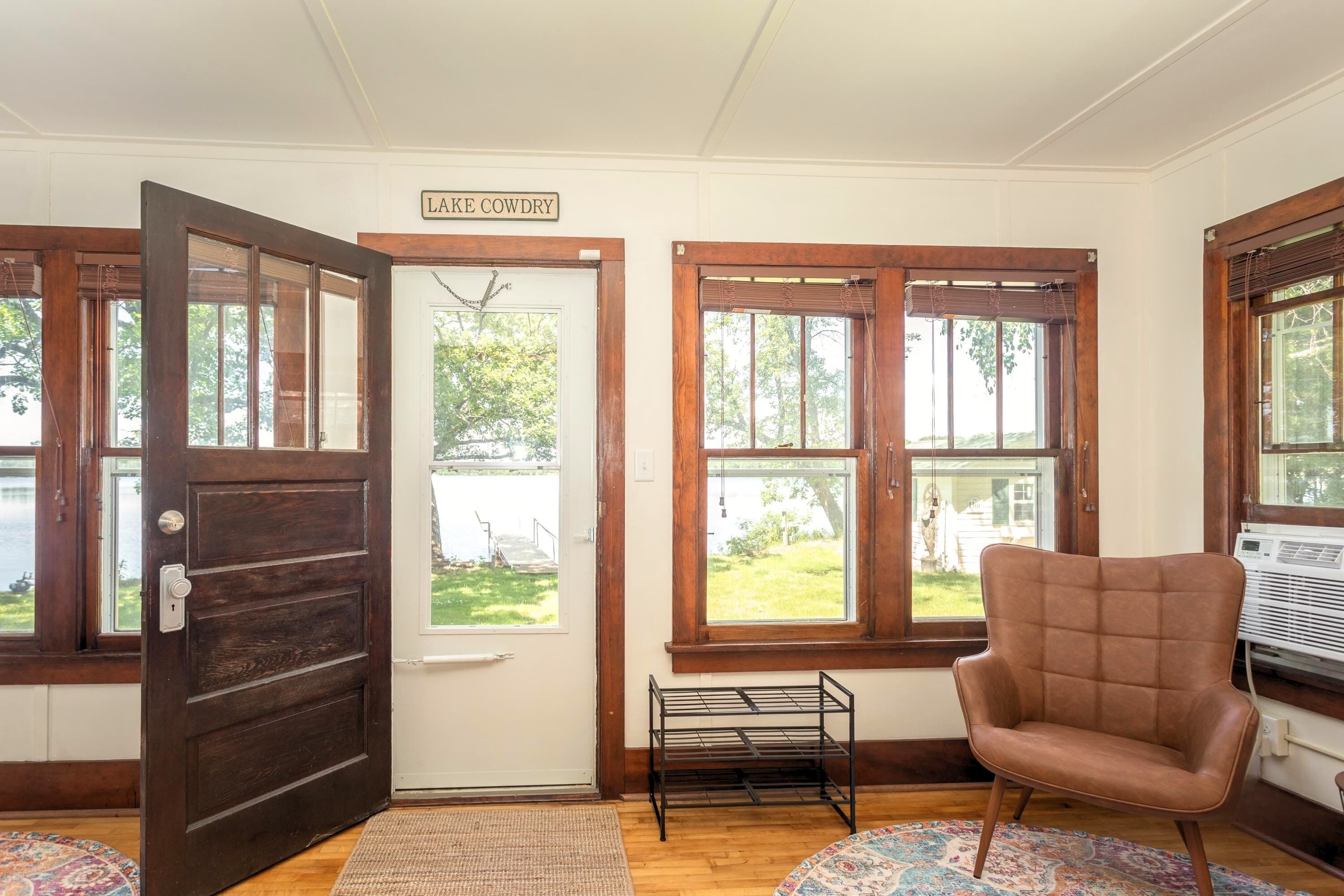Habitación con puerta de madera que dice "LAKE COWDRY", una silla con cojín marrón, un estante metálico pequeño y vista a un lago a través de amplias ventanas.