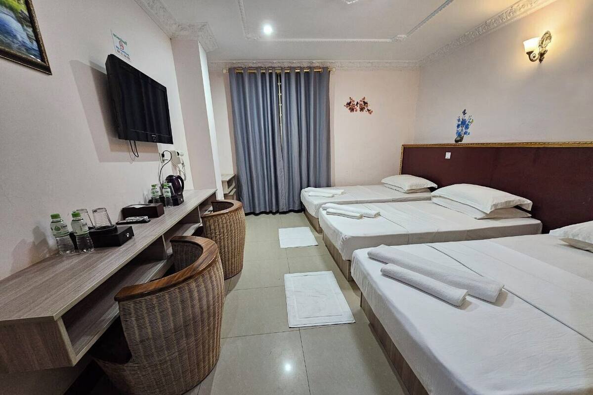 Ein Hotelzimmer mit drei Betten, einem an der Wand befestigten Fernseher, einem Schreibtisch mit Computer und Lampe sowie einem Fenster mit Vorhängen.