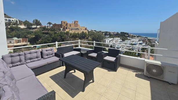 Eine Terrasse mit Gartenmöbeln, Blick aufs Meer und ein weit entferntes, schlossähnliches Gebäude.