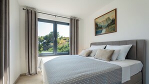 A bedroom with a large bed, a window with curtains, a painting on the wall, and a view of greenery outside. - Deluxe apartment Noeli (Krvavica)