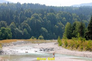 A river with people swimming and standing on the shore, surrounded by dense forest and hills.