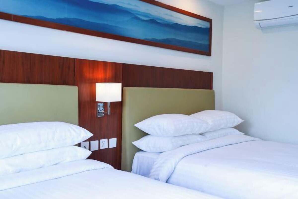 A hotel room with two beds, a nightstand with a lamp, a painting above the beds, and an air conditioner on the wall.