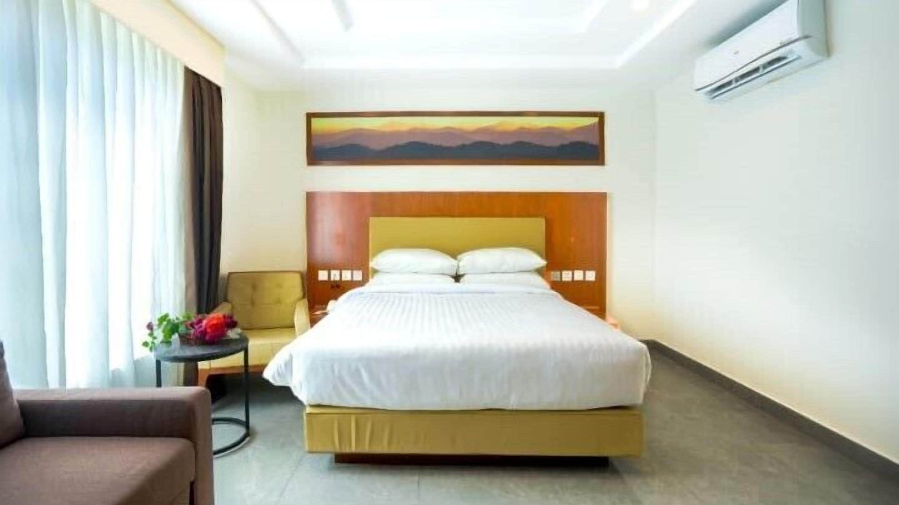 A hotel room with a bed, a chair, a small table with a plant, and a painting on the wall.