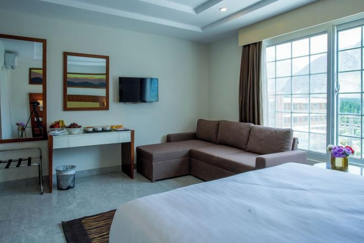 A hotel room with a bed, a desk with a TV, a sofa, and a window with a view.