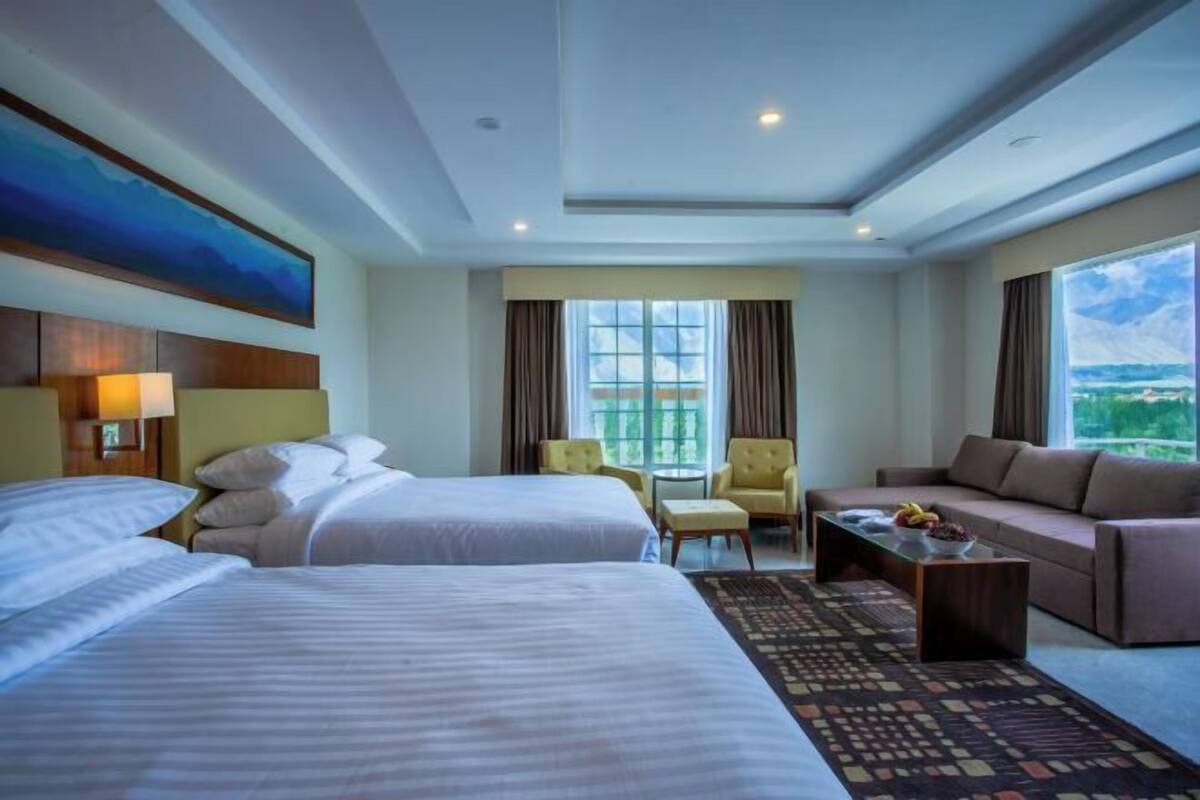 Habitación de hotel con dos camas, un sofá, una silla, una mesita y un ventanal grande con cortinas.