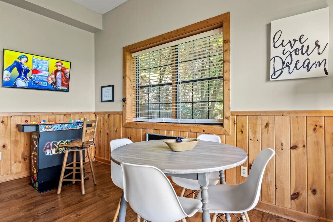 A room with a wooden paneled wall, a round table with chairs, a window with blinds, a television on the wall, and a motivational quote.
