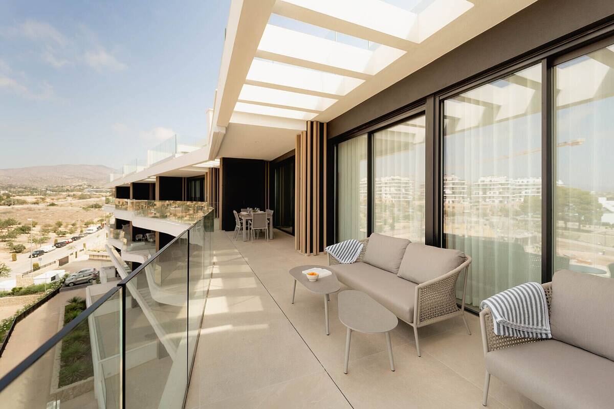 A modern balcony with a glass railing, a sofa, a small table, and a dining area with chairs.