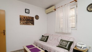 Una stanza piccola e ben illuminata con pavimento in legno, un divano rosa con cuscini decorativi, un piccolo tavolino con sottopiatti viola, una sveglia a parete, un quadro incorniciato, una finestra con persiane e un piccolo termosifone su un tavolino laterale.