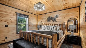 A wooden cabin bedroom with a large bed, a wooden headboard, a window, and a bear mural on the wall.